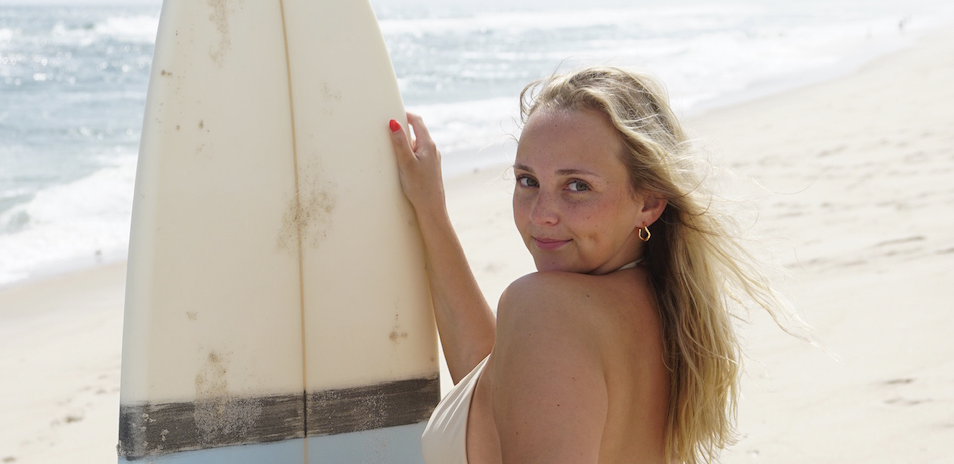 A woman looking over her shoulder beside her surfboard on the beach