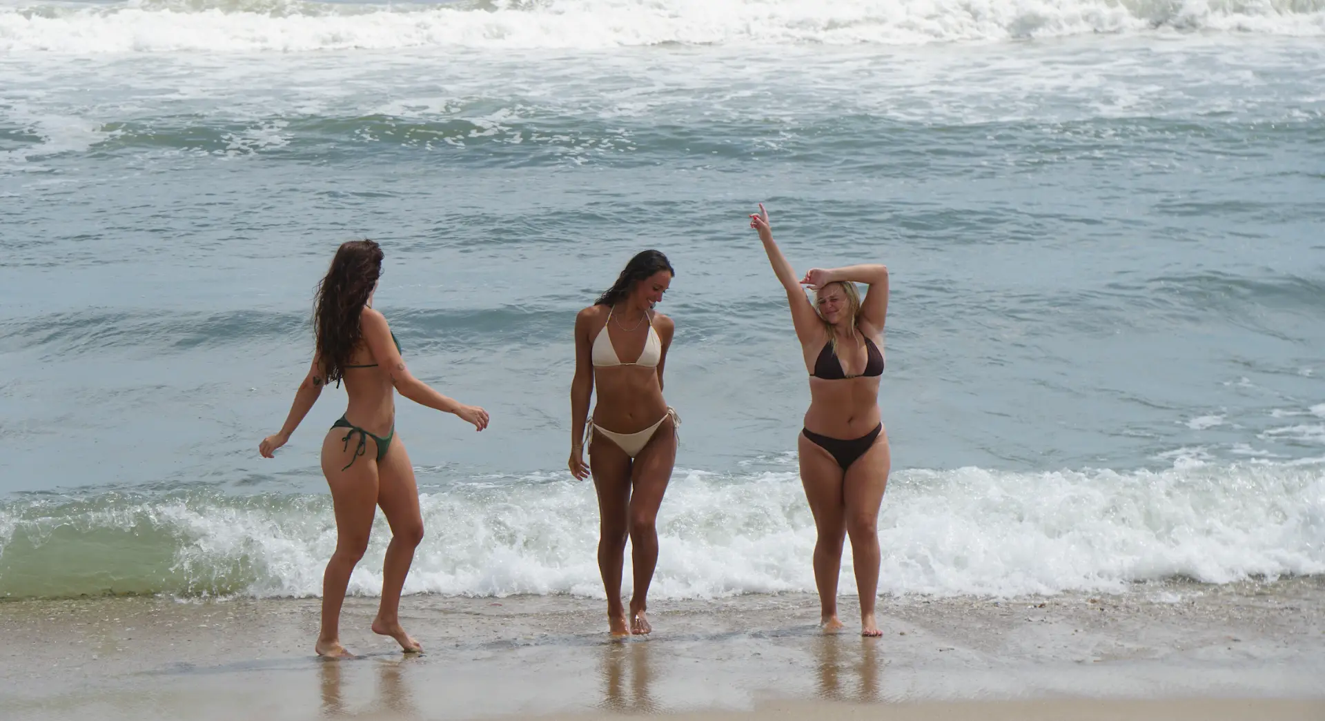 Three women in Olive, Coconut, and Espresso swimwear playing at the water's edge as waves roll in behind them